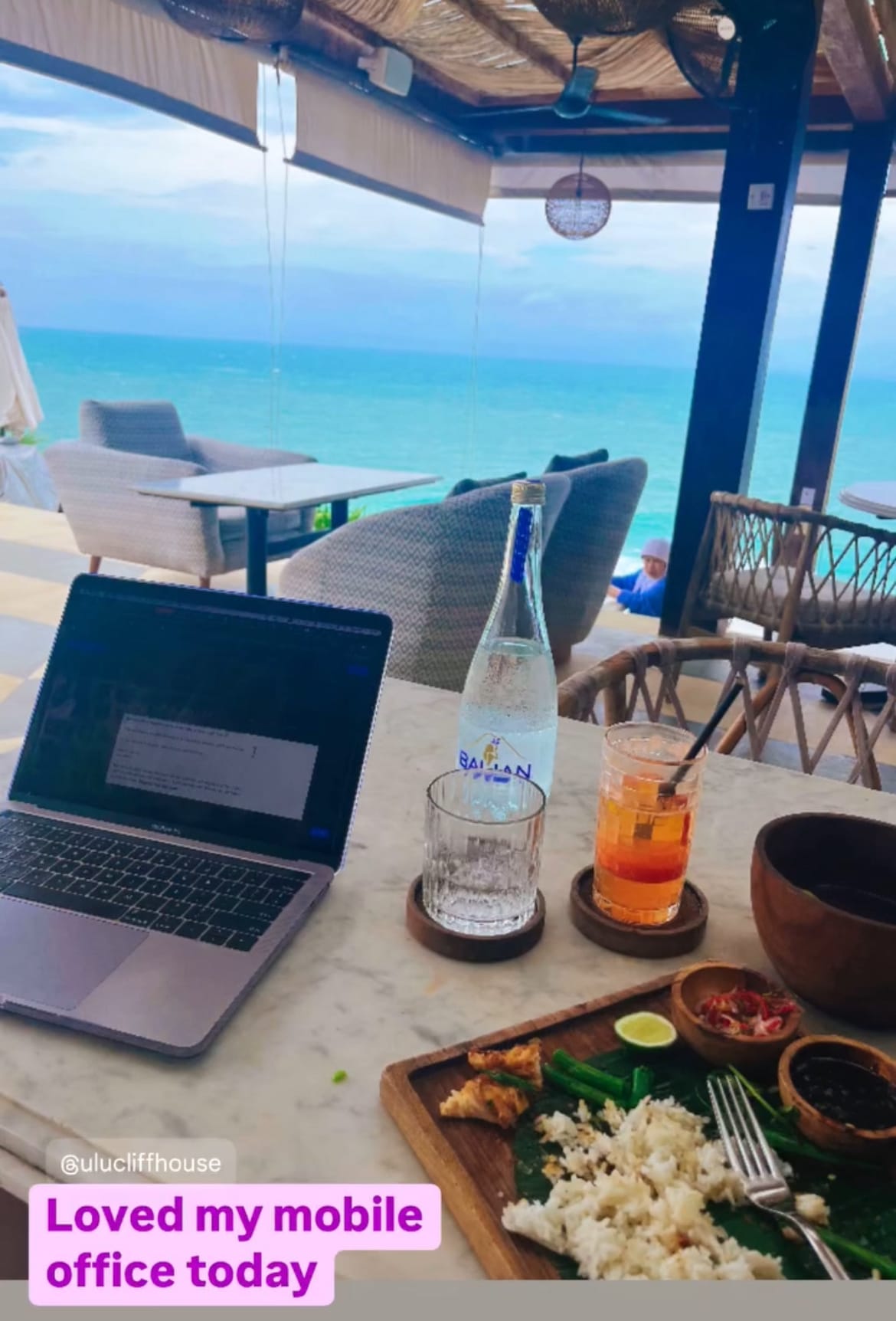 Laptop, sparkling water and a cocktail on a table at a clifftop bar in Bali, an afternoon work session with an ocean view