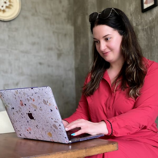 Marybeth Welty in a red dress working on her terrazzo-cased MacBook at a wooden table in an open-air Bali cafe