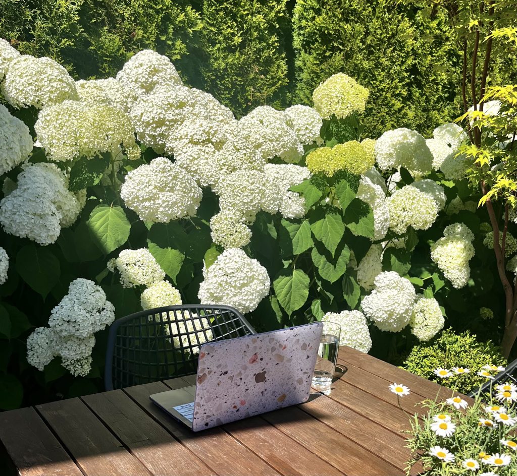 Open laptop in a terrazzo case on a wooden deck table surrounded by white hydrangeas, set up as an outdoor workspace